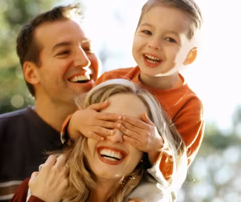 A woman smiling while a child on her shoulders covers her eyes, with a man smiling behind them