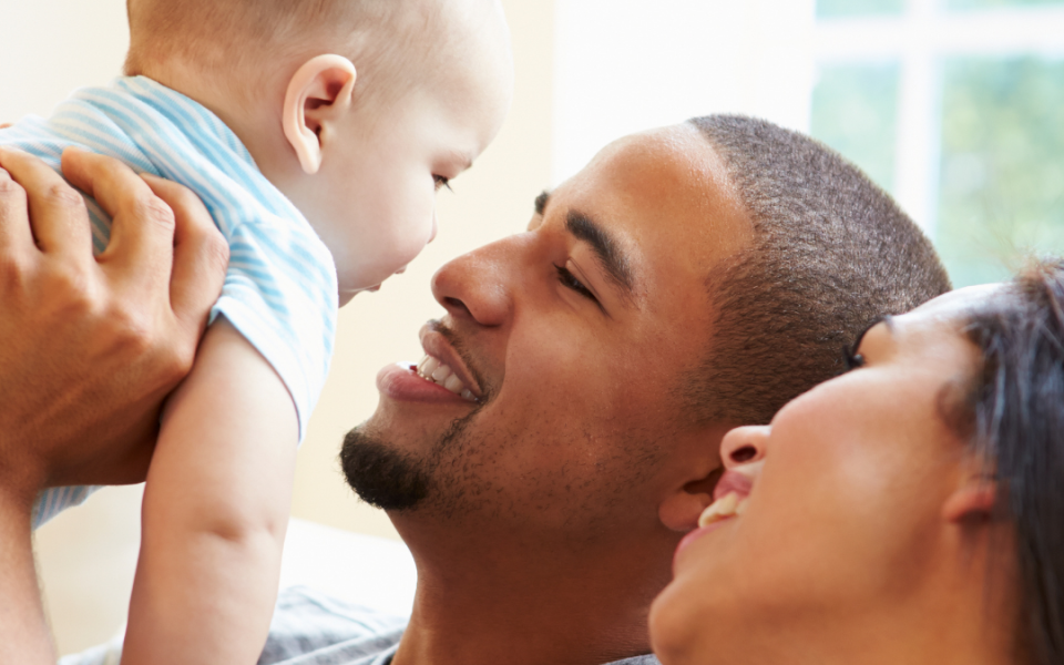 A man looking up at a baby in his hands, facing him, with a woman beside the man